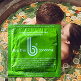 A couple embraces in a bathtub filled with fruit. In the foreground, there's a close-up of a green package labeled "bCondoms Ultra Thin Lubricated Condoms" from bCondoms, featuring product details and safety warnings in white text.